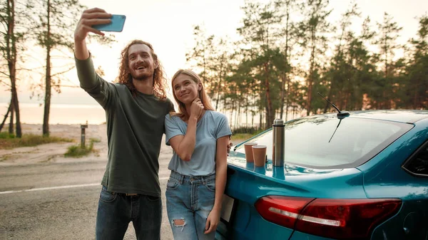 Hombre alegre usando teléfono inteligente mientras toma selfie junto con la atractiva mujer joven después de reparar el coche roto en el lado de la carretera — Foto de Stock