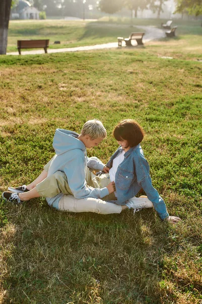 Feliz pareja lesbiana de chicas jóvenes sentadas en el césped en el parque de verano, mujeres enamoradas pasando tiempo juntas, sosteniéndose mutuamente —  Fotos de Stock