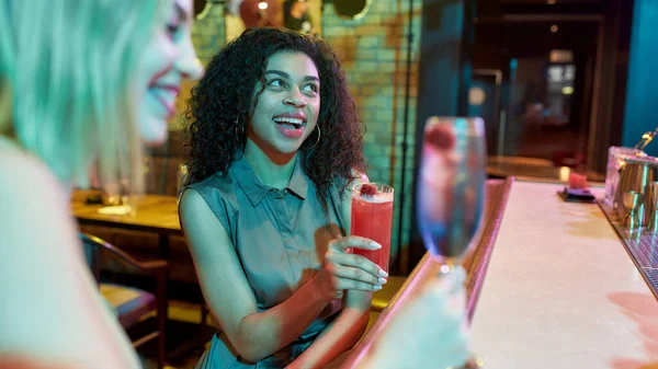 Dos mujeres divirtiéndose, brindando, bebiendo cócteles mientras están sentadas en el mostrador del bar. Amigos pasando tiempo en el club nocturno, restaurante — Foto de Stock