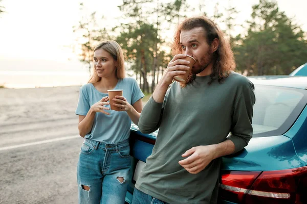 Jong stel, man en vrouw drinken hete koffie of thee na reparatie van de auto tijdens hun road trip — Stockfoto