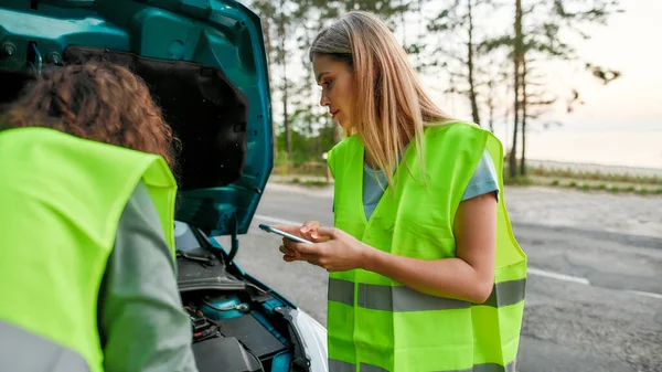 Couple wearing reflective vest having troubles with their auto, Young woman going to call car service or assistance while man looking under the hood of broken car
