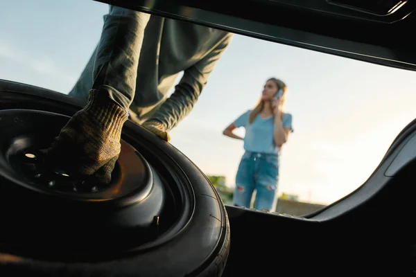 Primo piano delle mani maschili che tirano fuori la ruota di scorta, l'uomo sta per cambiare ruota dell'auto mentre aiuta la giovane donna con la sua auto rotta — Foto Stock