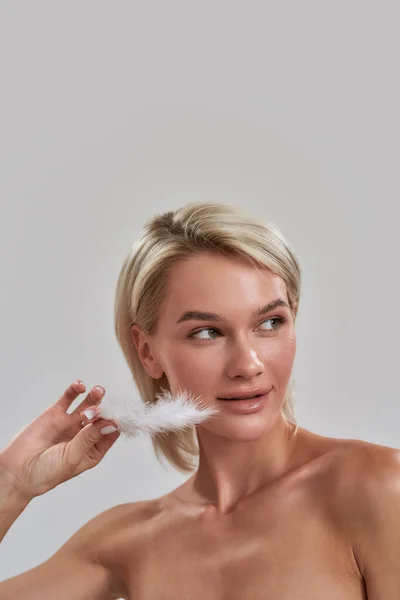 Retrato de una hermosa joven rubia con una piel fresca perfecta mirando a un lado, tocando su cara con pluma blanca mientras posa aislada sobre un fondo gris — Foto de Stock