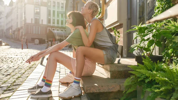 Jong lesbisch stel, twee meisjes in casual kleding en regenboog gekleurde sokken glimlachend, zittend op de trap tijdens een wandeling door de stad. LGBT, relatieconcept — Stockfoto
