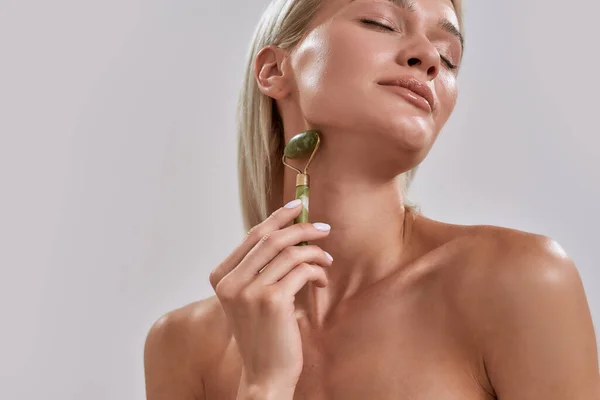 Retrato recortado de mujer joven con piel brillante perfecta usando rodillo facial de jade para el cuidado de la piel y el tratamiento de belleza mientras posa aislado sobre fondo gris — Foto de Stock