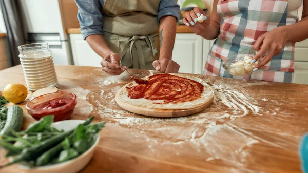 Ένα ζευγάρι να φτιάχνει πίτσα μαζί. Άντρας με ποδιά, με σάλτσα ντομάτας στη ζύμη, ενώ γυναίκα τον βοηθάει στην κουζίνα. Χόμπι, τρόπος ζωής — Φωτογραφία Αρχείου