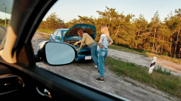 Meisje heeft problemen met haar auto, Man besloten om haar te helpen, hij kijkt onder de motorkap van een kapotte auto en probeert om het te repareren, Uitzicht vanuit een ander autoraam — Stockfoto