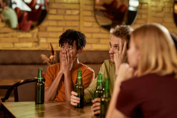 Un chico joven que se ve decepcionado mientras ve un partido de deportes en la televisión, bebiendo cerveza y animando al equipo en el bar. Concepto de personas, ocio y entretenimiento — Foto de Stock