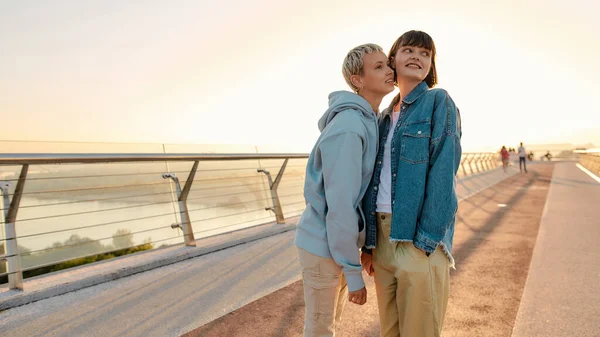 Feliz pareja lesbiana sonriendo, mirando a un lado mientras caminan en el puente y viendo el amanecer juntos —  Fotos de Stock