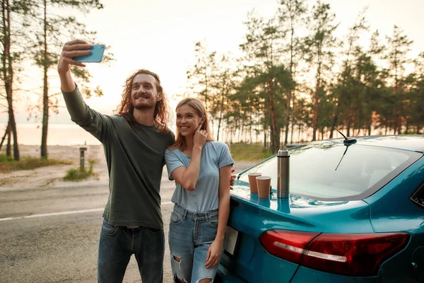 Hombre alegre usando teléfono inteligente mientras toma selfie junto con la atractiva mujer joven después de reparar el coche roto en el lado de la carretera — Foto de Stock