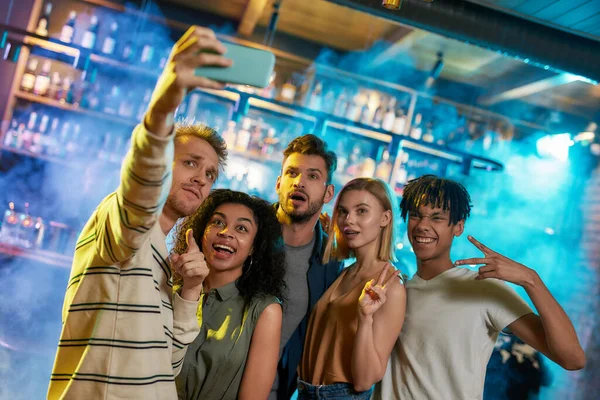 Fiatal férfiak és nők mosolyognak, miközben szelfit készítenek okostelefon használatával. Multiracionális baráti társaság bulizik a bárban. — Stock Fotó
