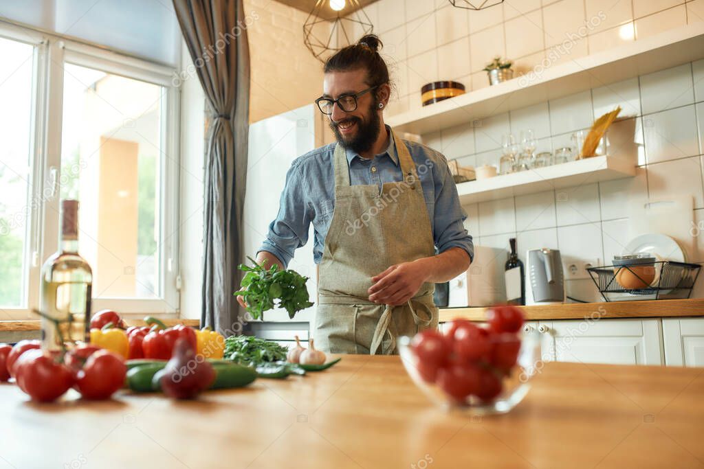 Cheerful young man, Italian cook in apron holding basil leaves while getting ready to prepare healthy meal with vegetables, standing in the kitchen. Cooking at home concept