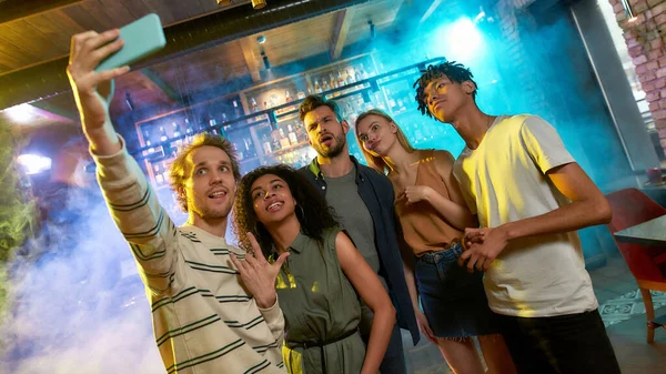 Hora de selfie. Hombres y mujeres jóvenes sonriendo mientras toman selfie usando un teléfono inteligente. Grupo multirracial de amigos pasando el rato en la fiesta en el bar — Foto de Stock