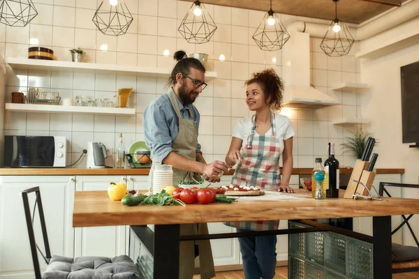 Pizza First. Young couple making pizza together at home. Man in apron adding mozzarella cheese on the dough while woman helping, looking at him. Hobby, lifestyle — Stock Photo, Image