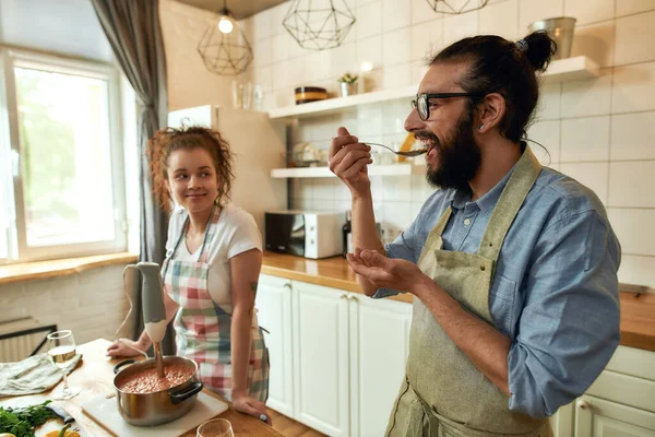 Uomo italiano, cuoco cucina zuppa degustazione mentre la sua ragazza lo guarda. Coppia allegra preparare un pasto insieme in cucina. Cucina casalinga, Cucina italiana — Foto Stock