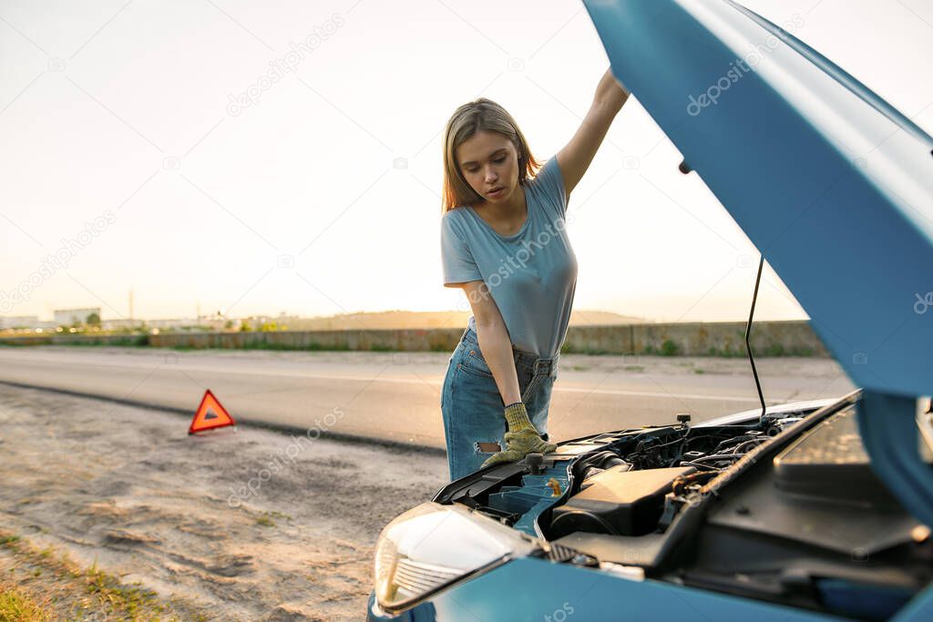 Close up shot of attractive young woman looking under the hood of her broken car while standing alone after car breakdown on the road side