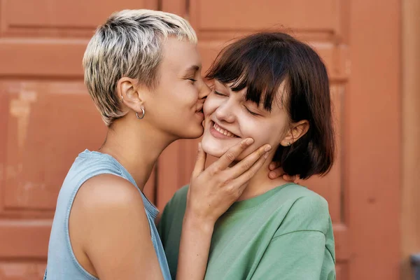 Pretty girl with short hair kissing her girlfriend outdoors. Young female couple smiling while standing in front of the door. LGBT, Sexual freedom concept — Stock Photo, Image