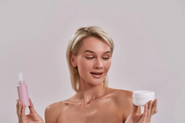 Retrato de una joven rubia con piel perfecta sosteniendo frasco y botella de diferentes productos cosméticos para el cuidado de la piel y eligiendo qué aplicar mientras posa aislada sobre fondo gris — Foto de Stock