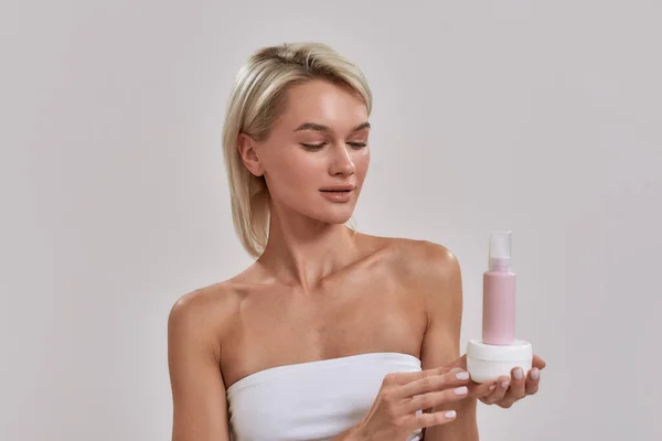 Retrato de mujer joven con piel brillante perfecta sosteniendo frasco y botella de diferentes productos cosméticos para el cuidado de la piel y mirándolo mientras posa aislado sobre fondo gris — Foto de Stock