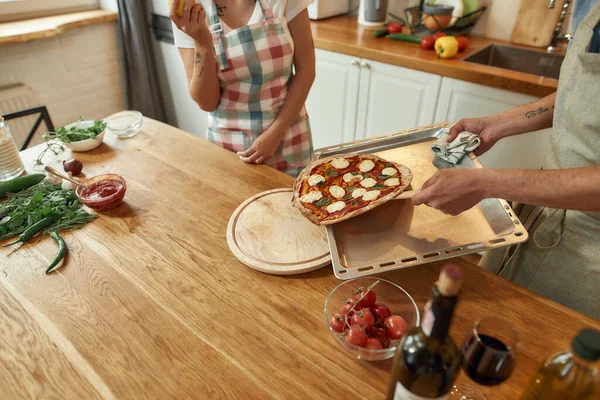 Recién salido del horno. Hombre joven en delantal, cocinar tomando bandeja con pizza recién horneada caliente fuera del horno. Una pareja haciendo pizza juntos. Hobby, estilo de vida — Foto de Stock