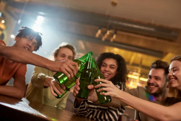 Un grupo de amigos felices en el bar tintinean botellas mientras beben cerveza juntos. Gente, ocio, amistad y entretenimiento — Foto de Stock