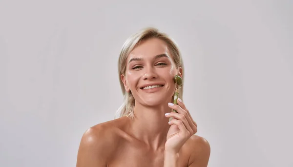 Portret młodej kobiety z idealną, świecącą skórą, uśmiechniętej do aparatu, używającej jadeitowego wałka do pielęgnacji skóry i zabiegu kosmetycznego, pozującej na szarym tle — Zdjęcie stockowe