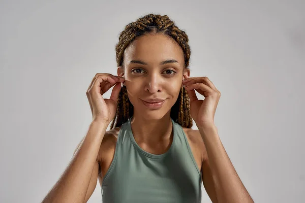 Portret van een jonge Afrikaanse fitnessvrouw met draadloze oordopjes, kijkend naar de camera en lachend staand geïsoleerd over een grijze achtergrond — Stockfoto