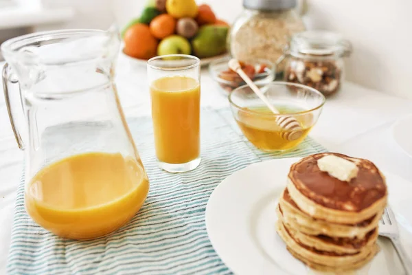 A jug and a glass of orange juice, honey in a bowl, a stack of pancakes and various fruits served for breakfast on the table — Stock Photo, Image