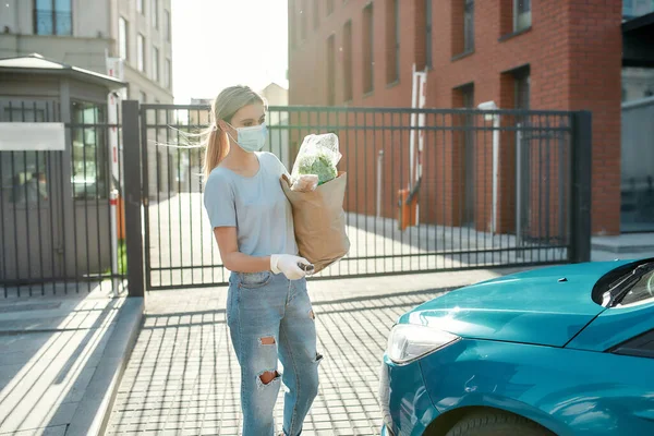 Jonge vrouw in medisch masker en beschermende handschoenen staan in de buurt van haar blauwe auto, ontgrendelen deur alarmsysteem terwijl het houden van boodschappentas — Stockfoto