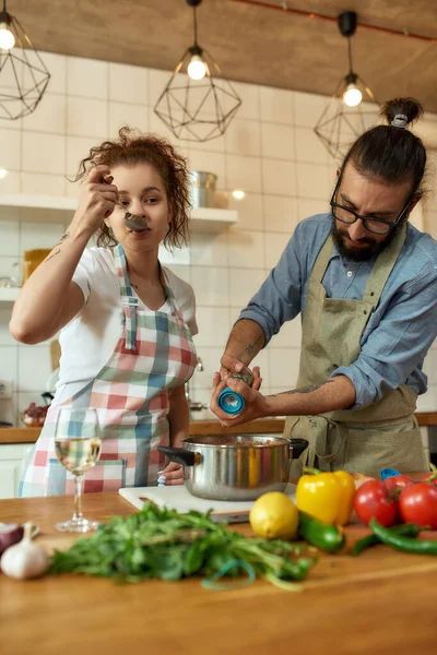 Italian man adding pepper, spice to the soup while woman tasting it. Couple preparing a meal together in the kitchen. Cooking at home, Italian cuisine