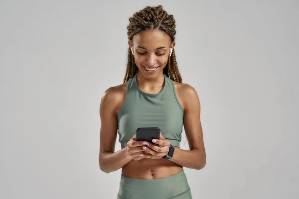 Giovane positiva mista gara fitness donna sms messaggio su smartphone e sorridente mentre in piedi in studio isolato su sfondo grigio, a riposo dopo l'allenamento — Foto Stock