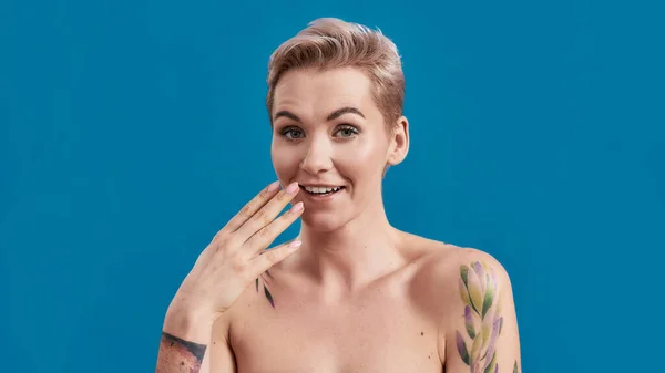 Scherzavo. Ritratto di bellezza di una giovane attraente donna mezza nuda tatuata con pelle perfetta guardando sorpreso dalla fotocamera con mano vicino alla bocca isolata su sfondo blu — Foto Stock