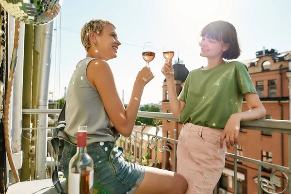 Две веселые девушки держат бокал вина, тосты во время вечеринки, празднуют на балконе. Молодая лесбийская пара проводит свой день вместе — стоковое фото