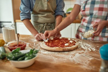 Dandik zevkler için. Evde birlikte pizza yapan bir çiftin görüntüsü. Önlüklü adam hamura domates sosu sürerken kadın mozzarella peyniri ekliyor. Hobi, yaşam tarzı