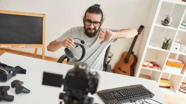 Man teknik bloggare som håller hörlurar och visar tummen ner medan du spelar in video blogg eller vlog om nya prylar hemma studio — Stockfoto