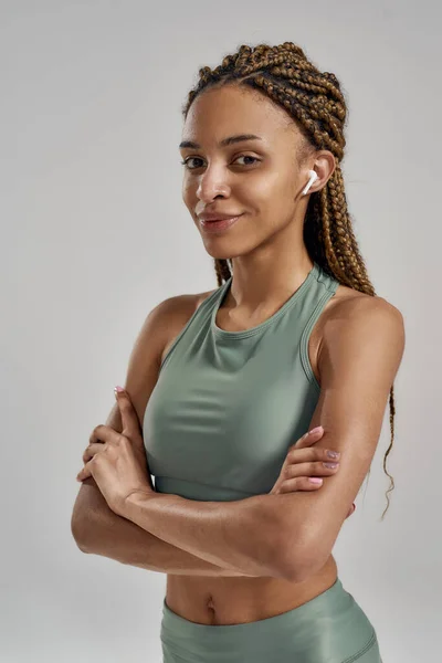 Scatto verticale di una giovane bella donna di fitness mista che indossa auricolari wireless guardando la fotocamera e sorridendo mentre posa isolata su sfondo grigio — Foto Stock