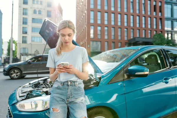 Ελκυστική νεαρή γυναίκα κρατώντας τηλέφωνο, καλώντας την υπηρεσία αυτοκινήτων έκτακτης ανάγκης, ενώ στέκεται, ακουμπώντας στο σπασμένο αυτοκίνητό της με ανοιχτή κουκούλα στο δρόμο της πόλης — Φωτογραφία Αρχείου