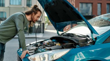 Odaklanmış görünen genç adam, şehir caddesinde dururken kırık arabasını açık kaputla inceliyor.