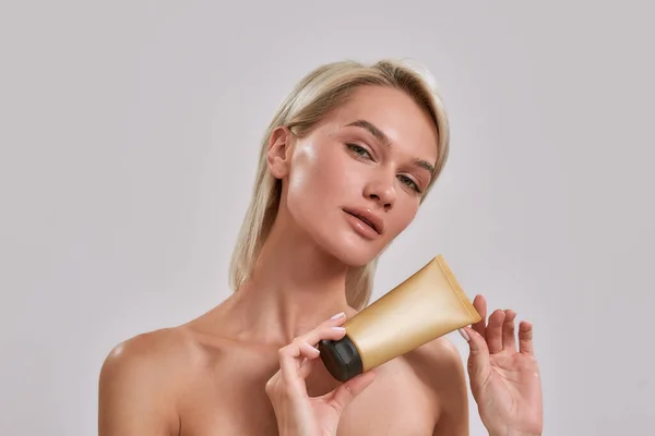 Retrato de mujer joven con una piel brillante perfecta mirando a la cámara, sosteniendo el producto de cuidado de la piel del protector solar mientras posa aislado sobre un fondo gris — Foto de Stock