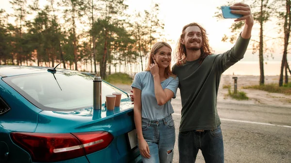 Hombre alegre usando teléfono inteligente mientras toma selfie junto con la atractiva mujer joven después de reparar el coche roto en el lado de la carretera — Foto de Stock