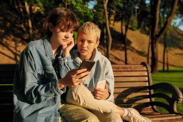 Δύο νεαρά κορίτσια να κρατάνε το κινητό τους, να κάθονται στον πάγκο στο πάρκο της πόλης. Ζευγάρι λεσβιών που χρησιμοποιούν τα τηλέφωνά τους ενώ περνούν χρόνο μαζί στην ύπαιθρο — Φωτογραφία Αρχείου