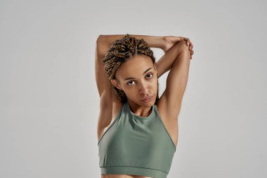 Isınma ve esneme hareketleri. Spor kıyafetli genç bir kadın spor salonunda spor yapmadan önce kollarını esnetiyor ve gri arka planda tek başına kameraya bakıyor.