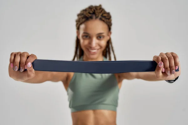 Fitness sakızıyla egzersiz yapıyorum. Spor kıyafetli genç bir kadın elastik bantla kameraya bakıyor ve gri arka planda tek başına dikilirken gülümsüyor. — Stok fotoğraf
