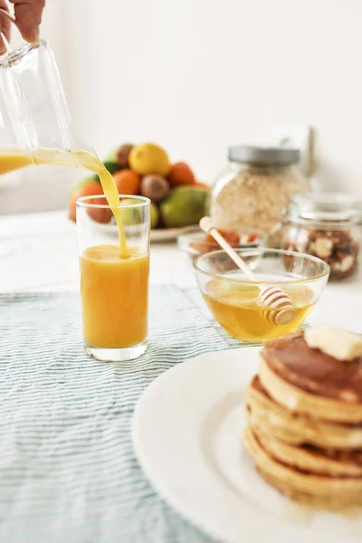 The process of pouring orange juice in the glass, Honey in a bowl with dipper, pancakes and various fruits served for breakfast on the table — Stock Photo, Image