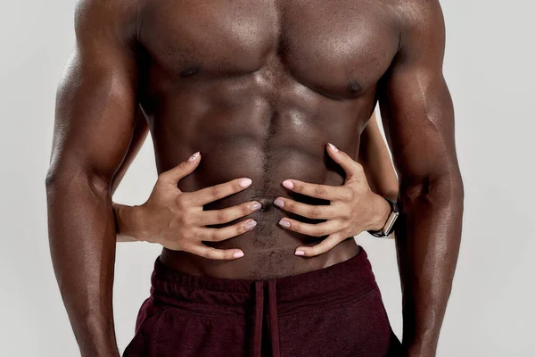 Perfecte buik. Vrouwelijke handen omhelzen, raken gespierde Afro-Amerikaanse man buikspieren geïsoleerd over grijze achtergrond. Sport, workout, bodybuilding concept — Stockfoto