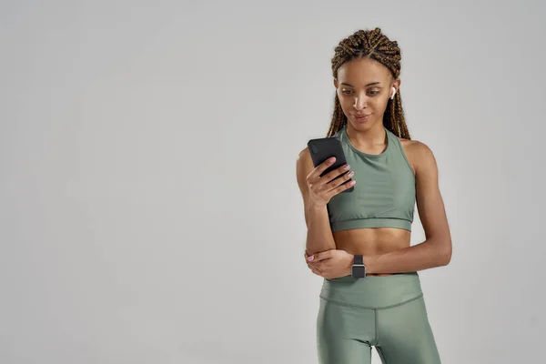 Joven mujer fitness de raza mixta con auriculares inalámbricos usando su teléfono inteligente después del entrenamiento mientras está de pie en el estudio aislado sobre fondo gris — Foto de Stock