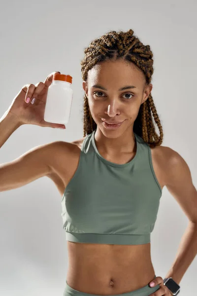 Colpo verticale di una giovane donna mista in abbigliamento sportivo bottiglia con integratori nutrizionali fitness mentre in piedi isolato su sfondo grigio — Foto Stock