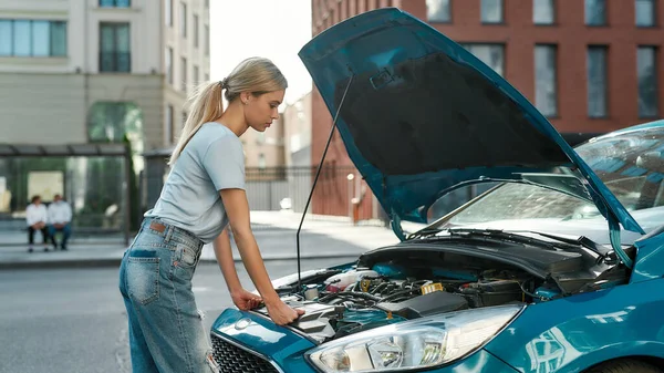 Aantrekkelijke jonge vrouw onderzoeken, kijken naar haar kapotte auto met open motorkap terwijl staan op de straat — Stockfoto