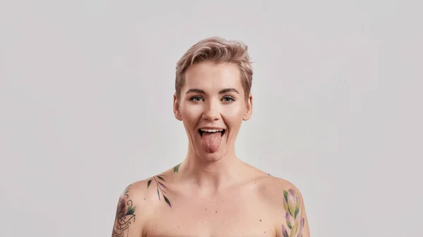 Retrato de una joven atractiva mujer semidesnuda tatuada con la piel perfecta mostrando su lengua a la cámara aislada sobre fondo claro — Foto de Stock