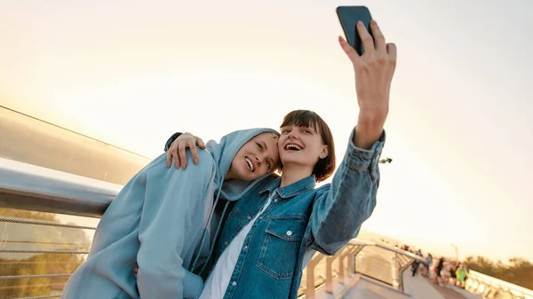 Pareja de lesbianas de pie en el puente, sonriendo mientras toman una foto selfie, viendo el amanecer juntos —  Fotos de Stock